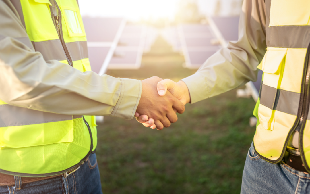 A handshake between two people in high-vis jackets is in the foreground, a solar farm is behind them.