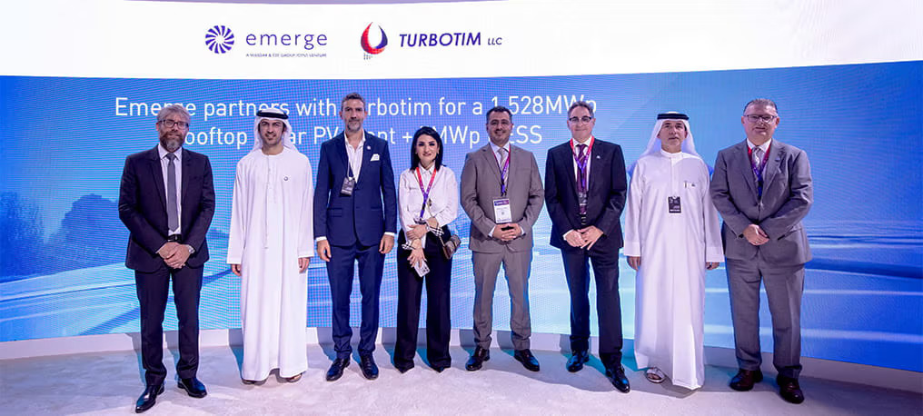 People stand in front of a sign with company logos for "Emerge" and "Turbotim"